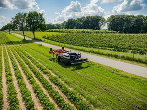Fahrerloser Agrarroboter mit Raupenketten und einem Anbaugerät zum Pflügen fährt am Feldrand entlang. Daneben verläuft eine Straße. 