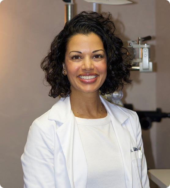 Dr. Kalindi Desai, Williamsville optometrist, smiling in a white lab coat with eye exam equipment in the background.